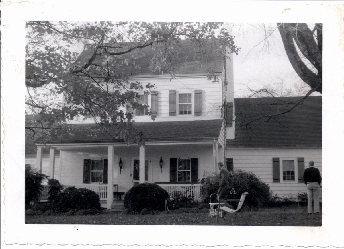 Black and white photograph with a scalloped border showing a two-storey white house with a full-width porch and a person standing at the right.