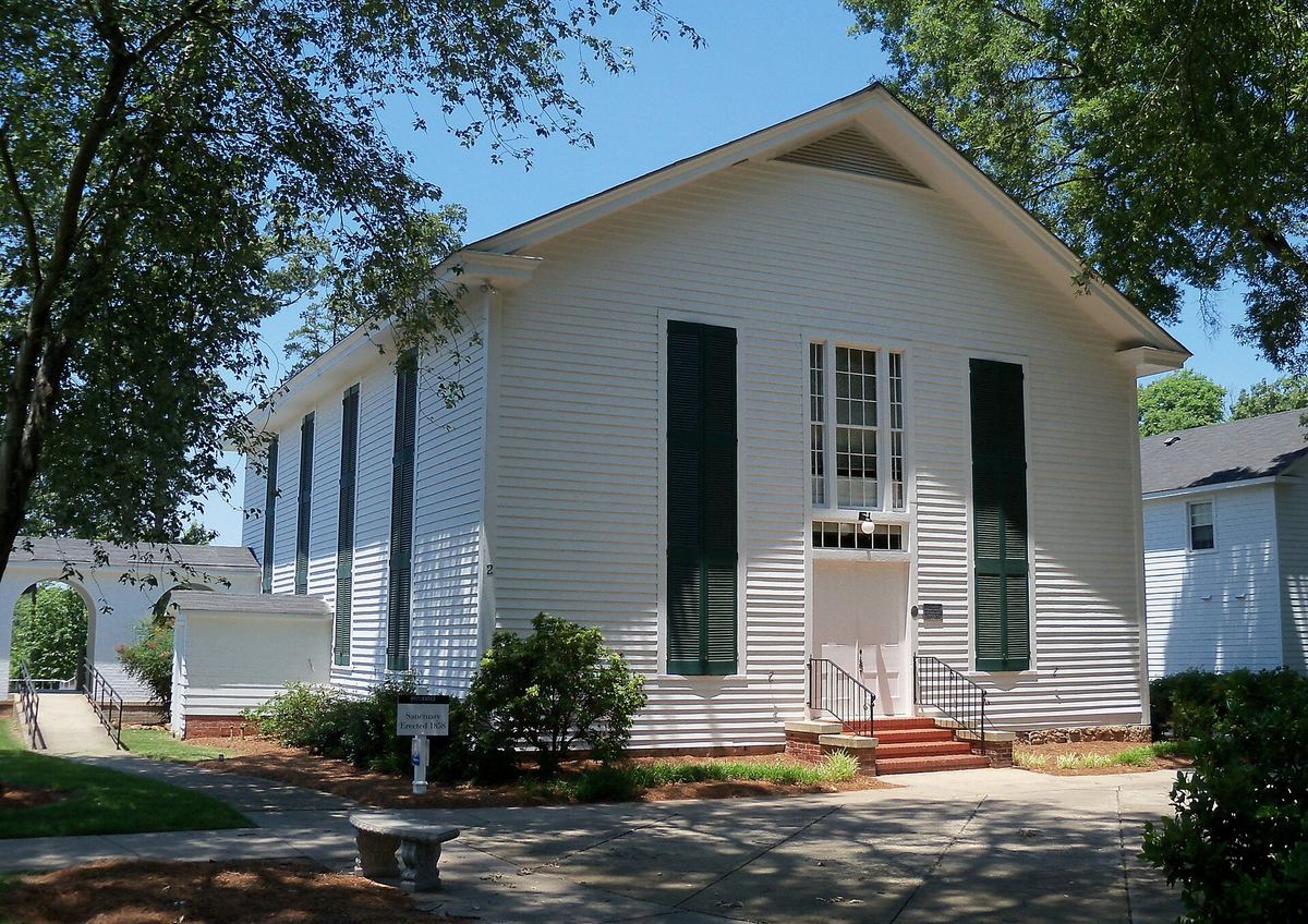 Small white clapboard church with dark green shutters, a gabled front and steps up to the entrance, among trees.