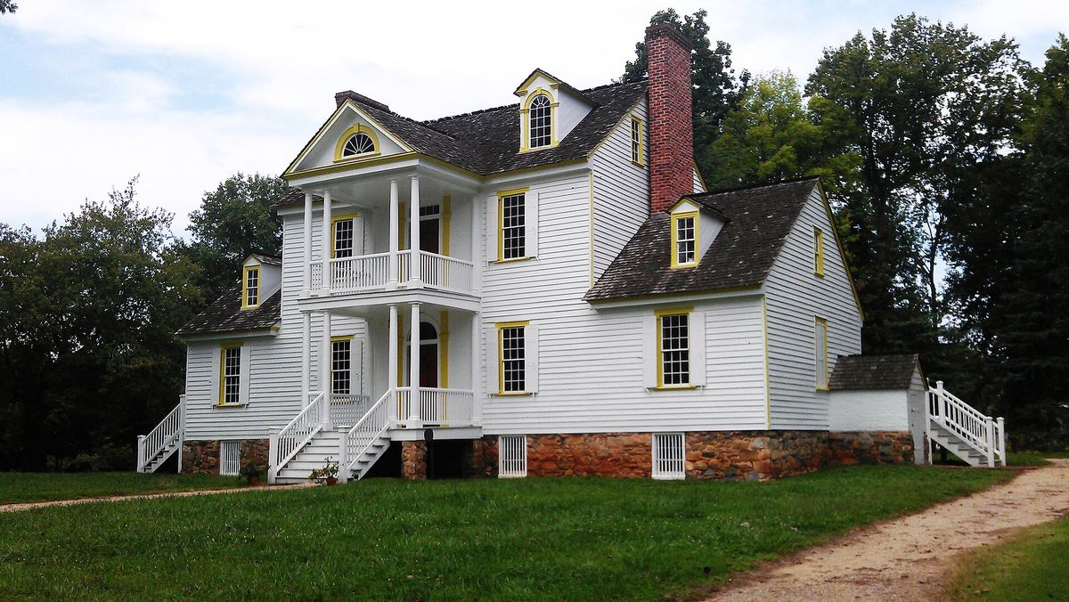 Two-storey white clapboard house with a central two-tier porch, tall red brick chimney and dormer windows, on an open lawn.