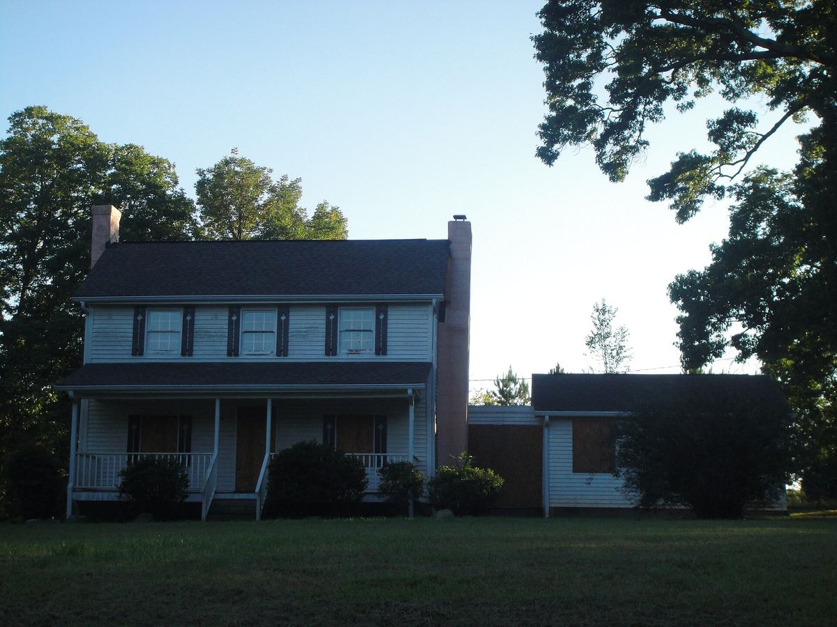 Two-storey house with dark siding and a covered porch, with a lower modern extension to the right, among tall pines.