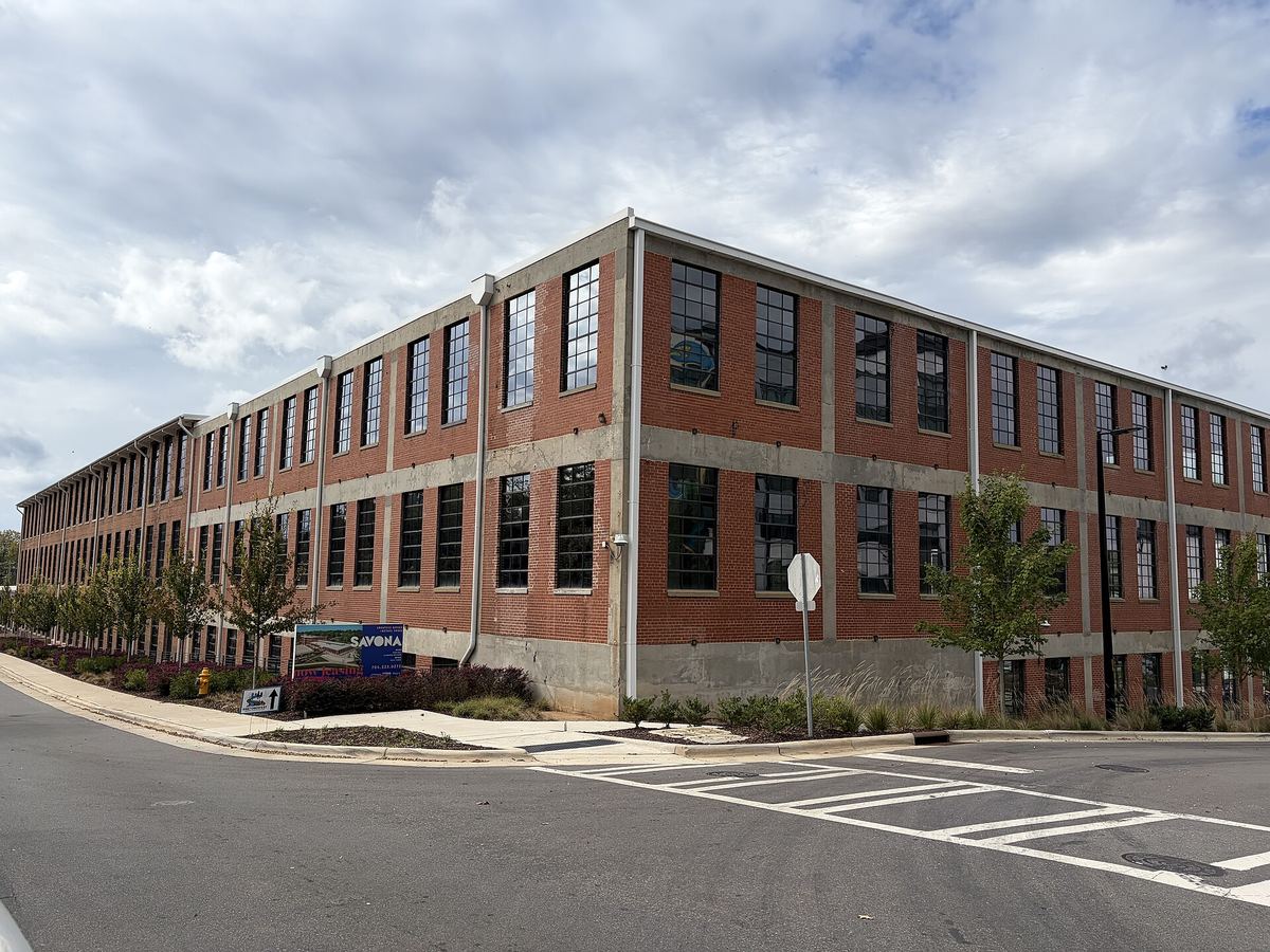 Long three-storey red brick mill building with regular banded windows, seen from a road junction with a pedestrian crossing in the foreground.