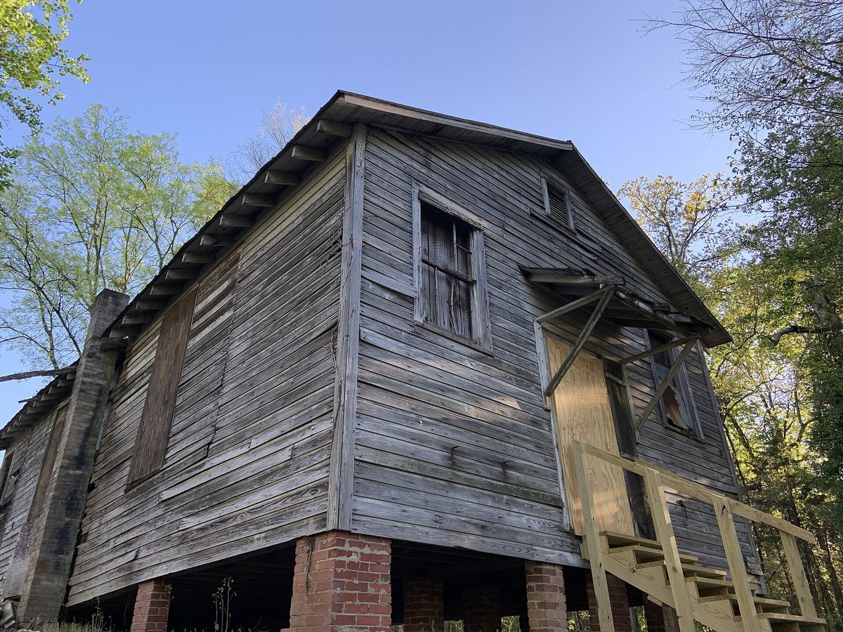 Weathered bare timber schoolhouse with missing boards and a collapsing roofline, raised on brick piers among trees.