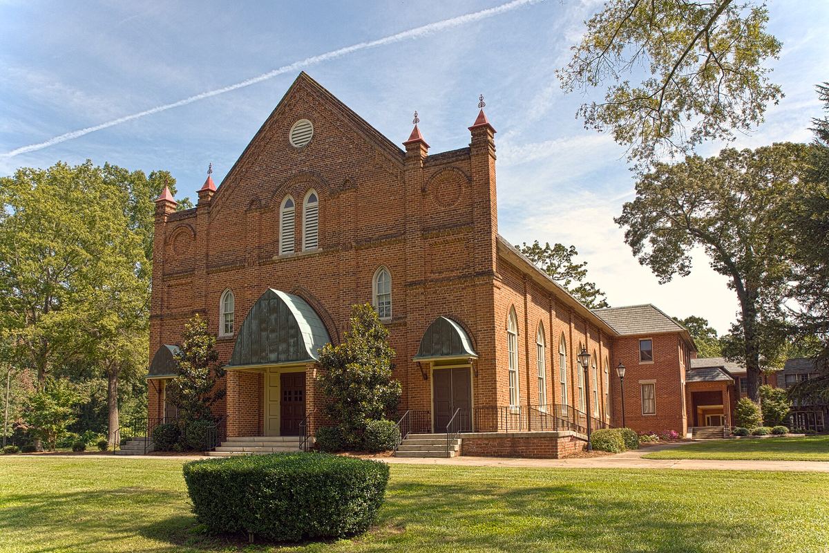 Red brick church with a large arched entrance recess, round-headed windows and twin pinnacles flanking the gable, on a mown lawn.