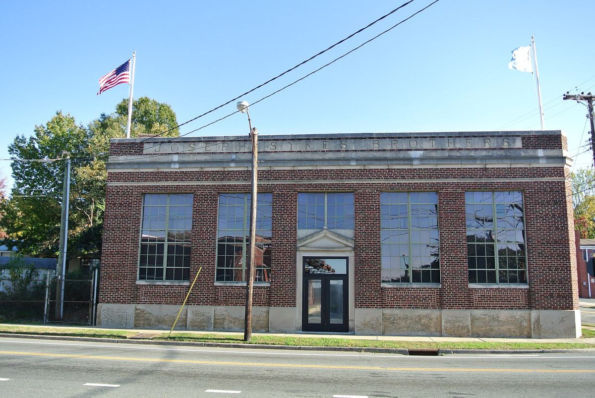 Single-storey red brick commercial building with tall multi-paned windows and a stone parapet, two flagpoles above, on a wide empty street.