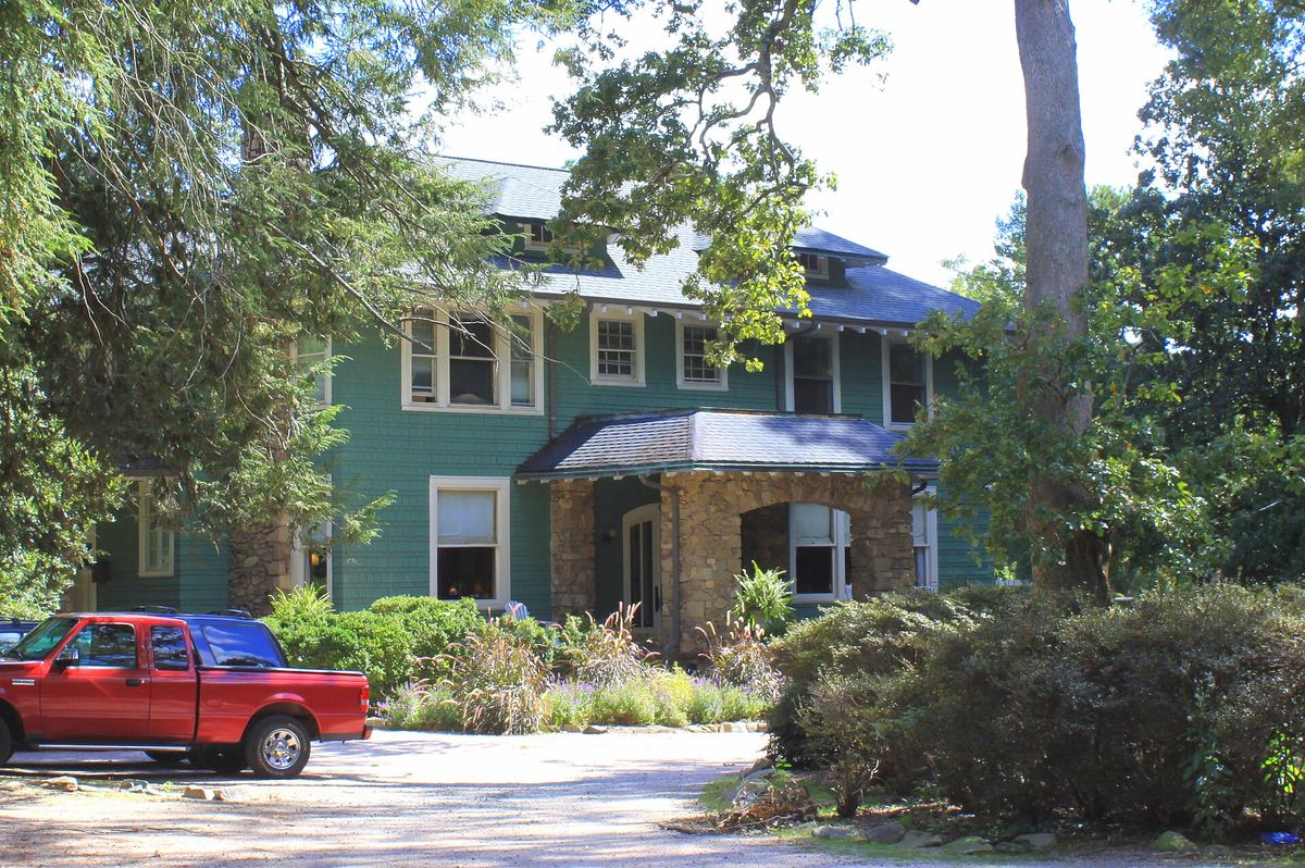 Green-painted timber house with cream trim and a low-pitched roof, largely screened by mature trees and shrubs, with a red pickup truck parked outside.