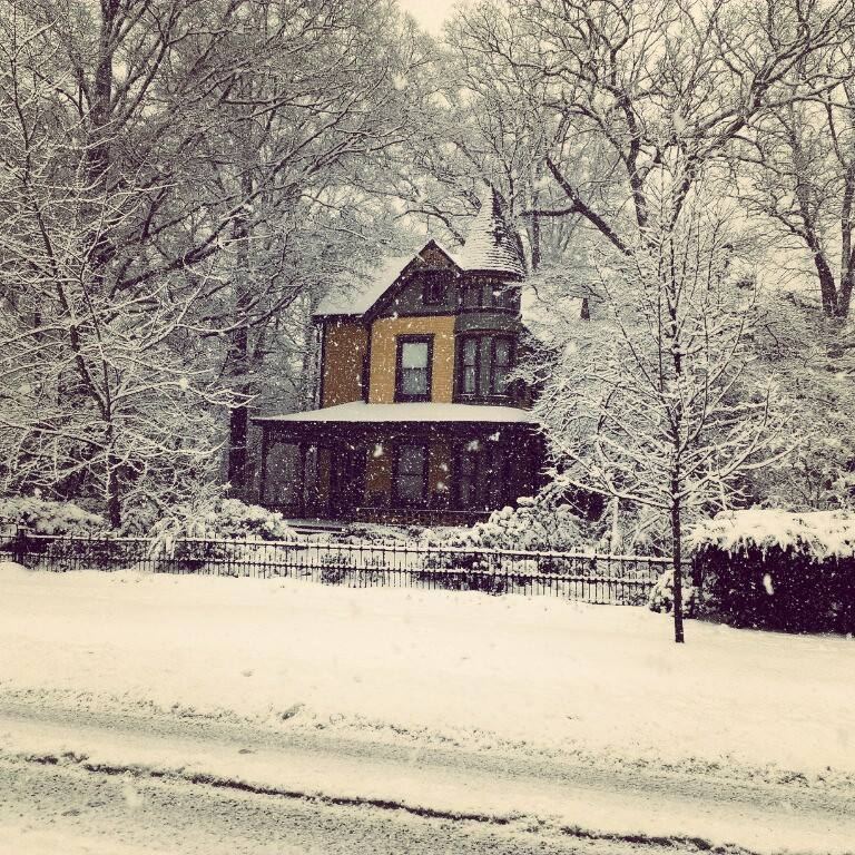 A Victorian house with a conical-roofed corner turret, steep gables and a deep wrap-around porch, seen across a snow-covered lawn and road during falling snow. Bare trees heavy with snow surround it and an iron railing runs along the front boundary.
