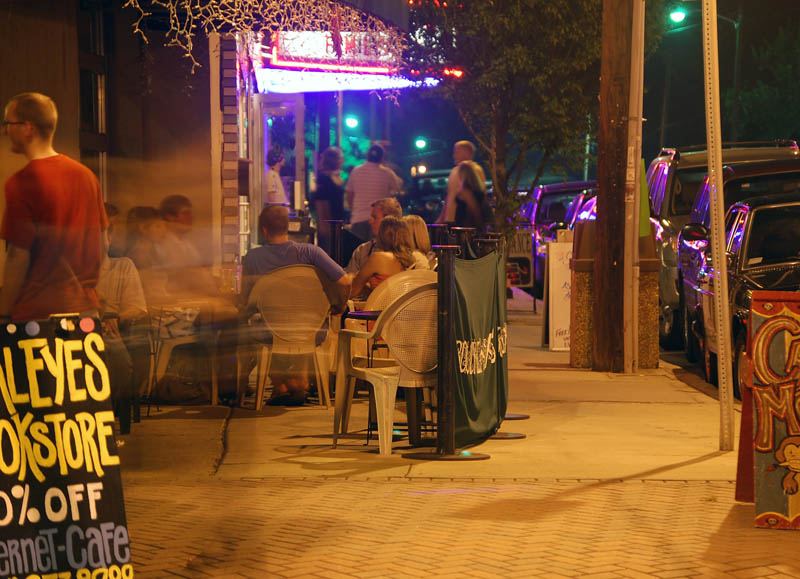 Evening scene outside a bar with people seated at outdoor tables under coloured lights, and a lit stage area visible through the opening.