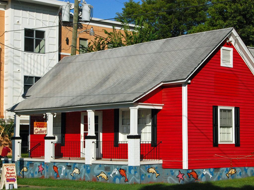 A small bright red weatherboard cottage with a grey shingle roof, white porch posts and black shutters. A hand-painted sign on the porch reads 'Studio True Gallery', a sandwich board at the kerb reads 'Now open', and the foundation wall is painted with a mural of fish and starfish on a blue-grey ground.