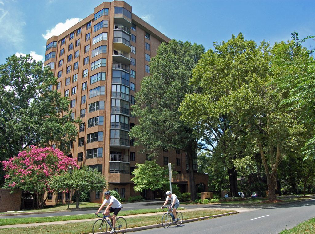 Mid-rise brick and glass apartment block behind mature trees, with two cyclists riding along the road in the foreground.