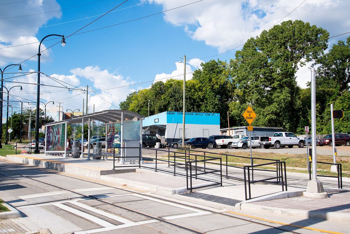 Modern light rail or bus stop with a glass shelter and metal railings on a new concrete platform, with parked cars and trees behind.