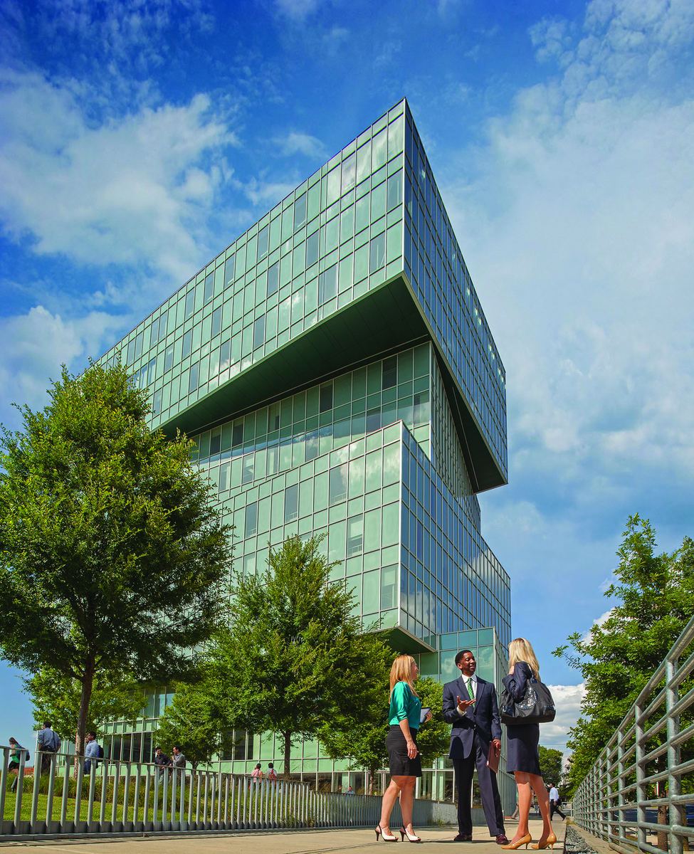 A green-tinted glass office tower whose upper block projects outward over the floors below, photographed from below against a blue sky with clouds. Trees and a metal railing line the pavement, where three people in business dress stand talking.