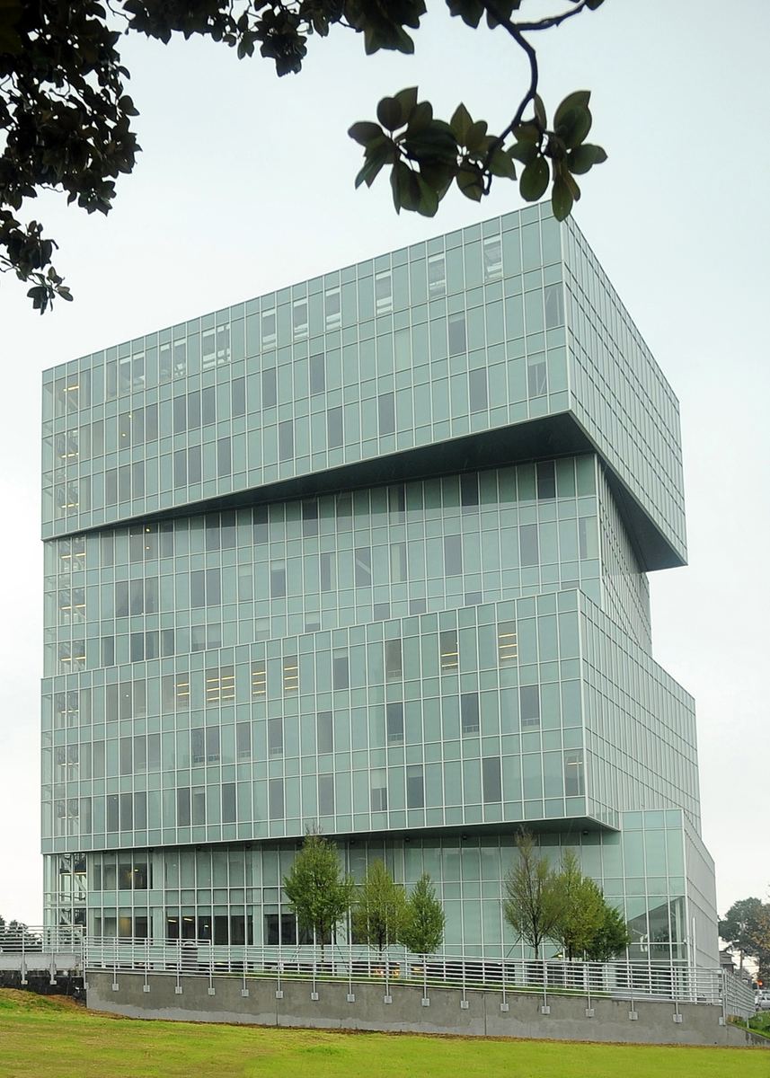A green-tinted glass tower of about ten storeys, its upper section cantilevered out over the lower floors, seen from a grass bank under a pale overcast sky with magnolia branches framing the top of the frame. A metal railing and young trees run along the base.