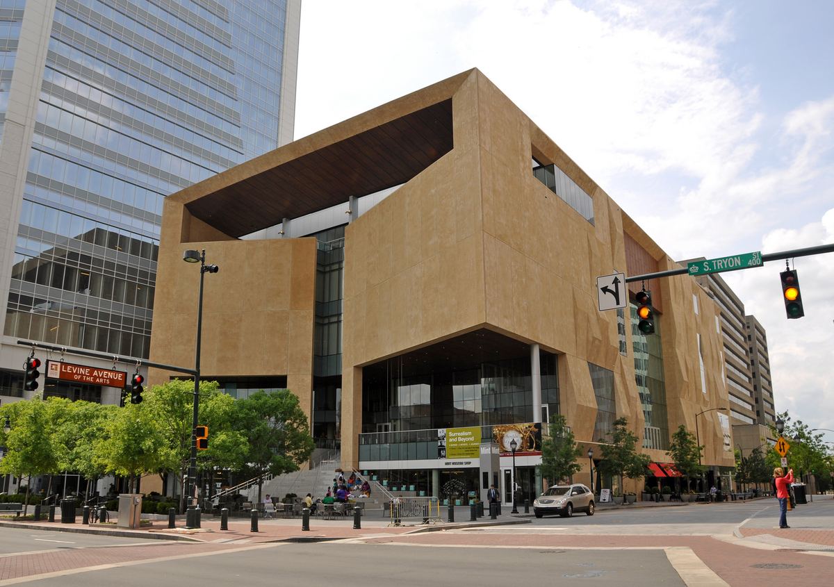 An angular building faced in pale sand-coloured panels with sharply folded planes and a deep overhanging upper storey, standing on a street corner beside a glass office tower. A green street sign reads 'S. Tryon St 400', a banner on a lamp post reads 'Levine Avenue of the Arts', and a banner on the building advertises an exhibition.