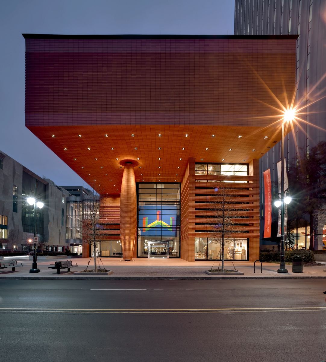 Museum building at night with a deep terracotta-coloured overhanging upper storey supported on a single central column, lit from beneath.