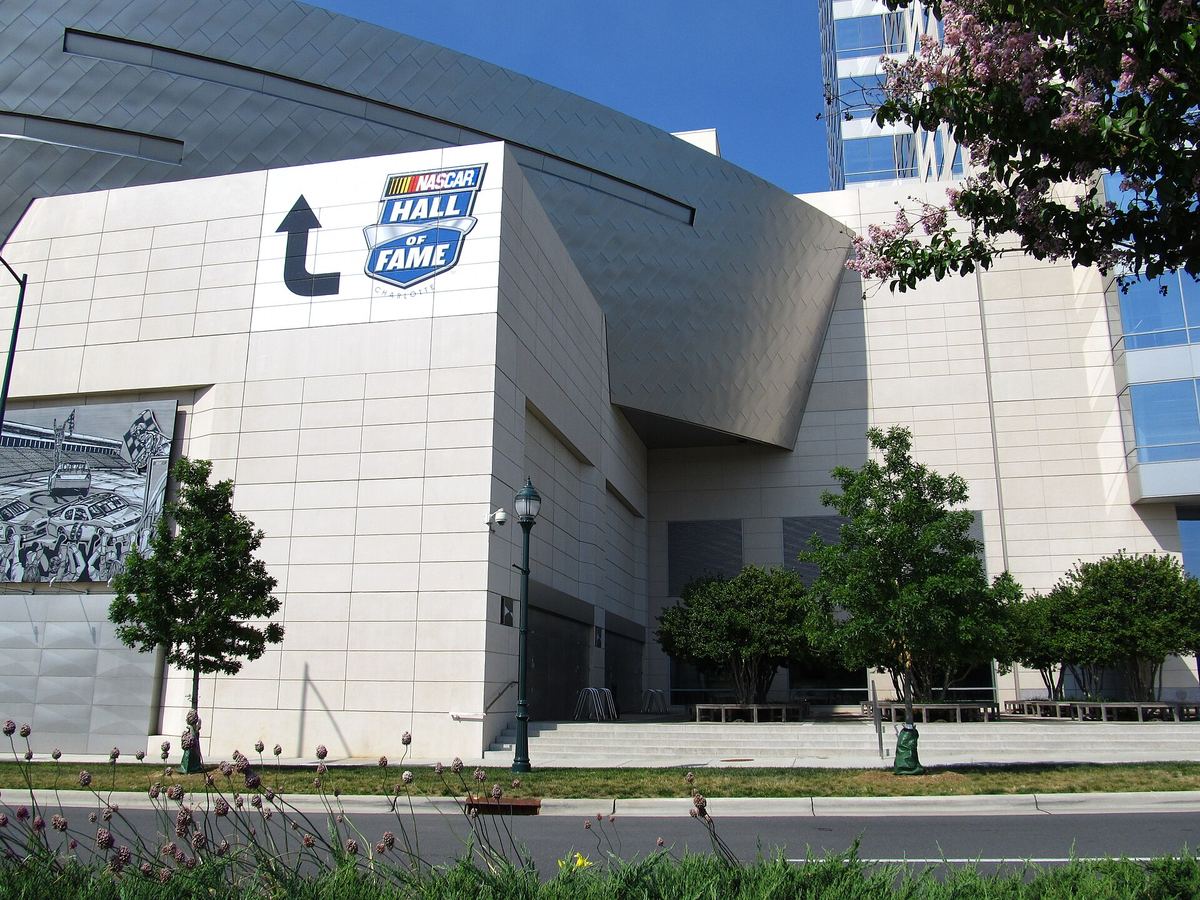 Curved white and silver museum building with a large banner reading NASCAR Hall of Fame, beside a landscaped pavement.