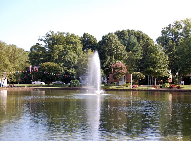 A lake with a tall central fountain plume, surrounded by mature trees in early autumn colour, with bunting strung along the far bank.