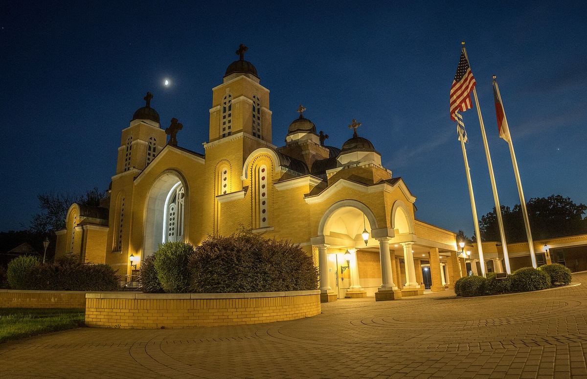 Pale stone Orthodox cathedral lit at night, with domed towers, an arched entrance portico and three flags on poles to the right.