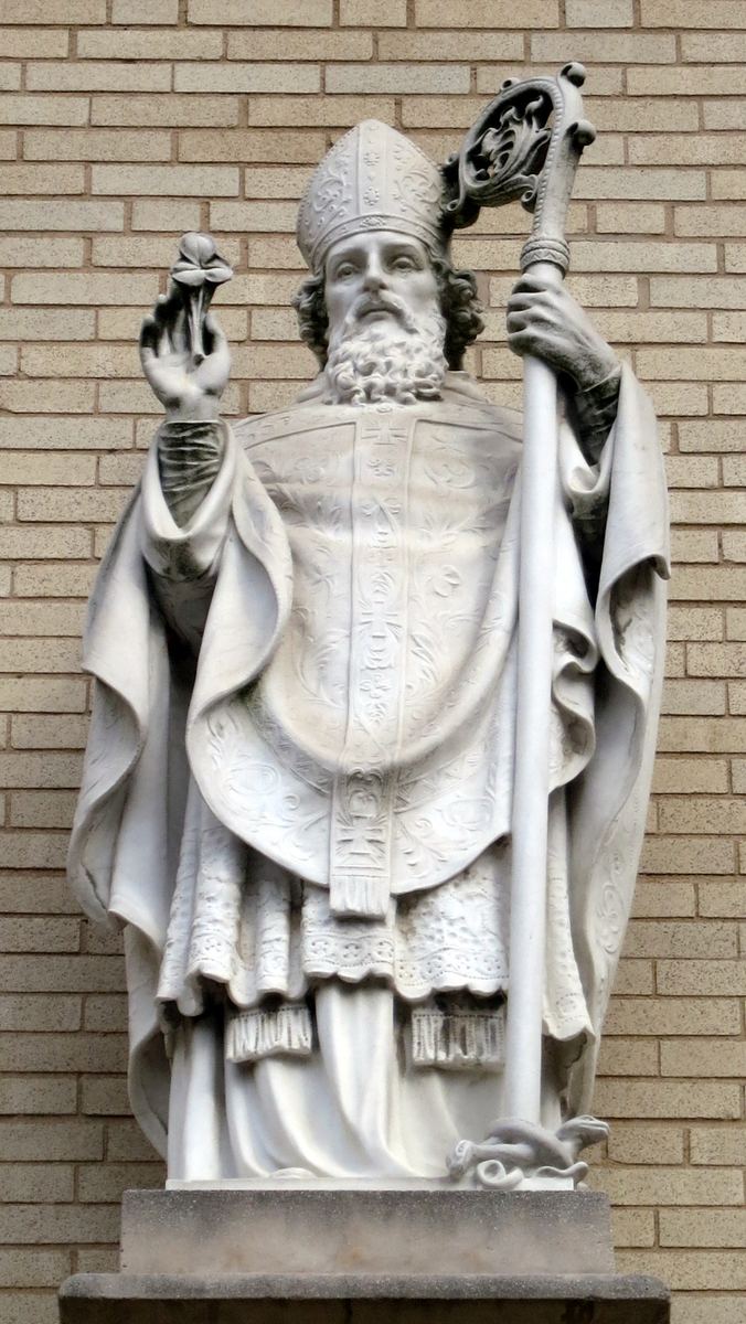 A white marble statue of a bearded bishop standing against a buff brick wall on a grey stone plinth. He wears a mitre, an embroidered chasuble and a flowing cope, holds a crozier in his left hand and raises a shamrock in his right, with a serpent carved at his feet.