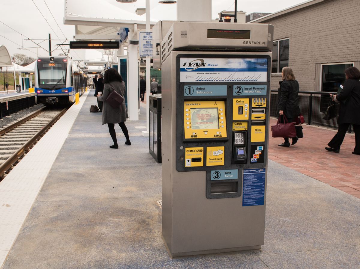 A light rail ticket machine on a station platform, with a blue and white tram at the platform edge and passengers walking past.