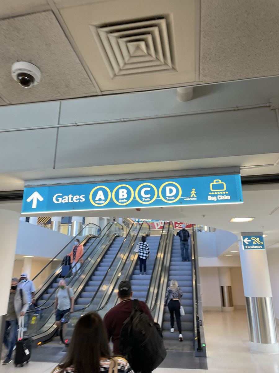 Airport concourse interior with escalators and an overhead blue sign reading Gates A B C D with an arrow, and travellers with luggage.
