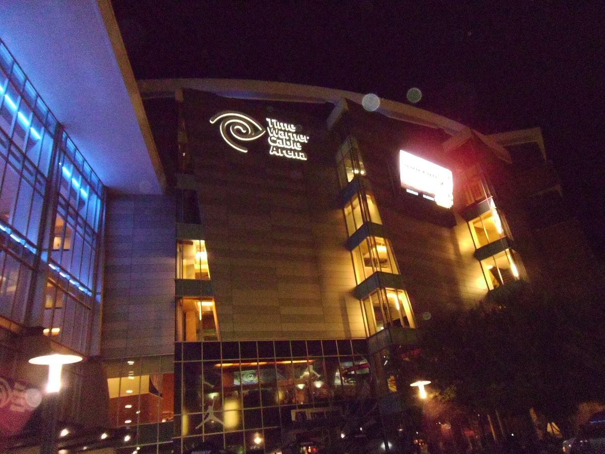Illuminated arena entrance at night with a curved glass and stone facade and a lit logo sign on the upper wall.