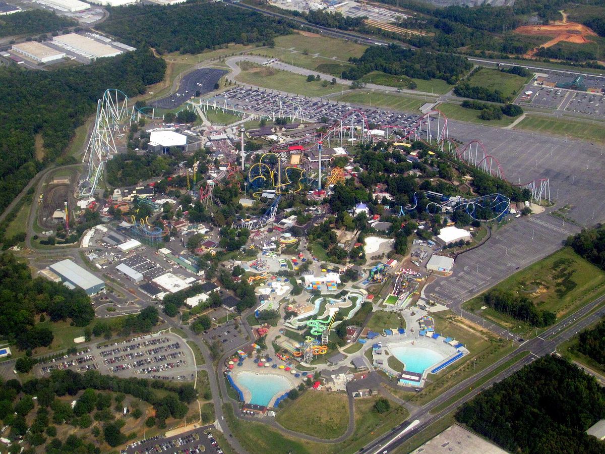 Aerial view of a large theme park with rollercoasters, a water park with blue pools, and extensive car parks surrounded by woodland.