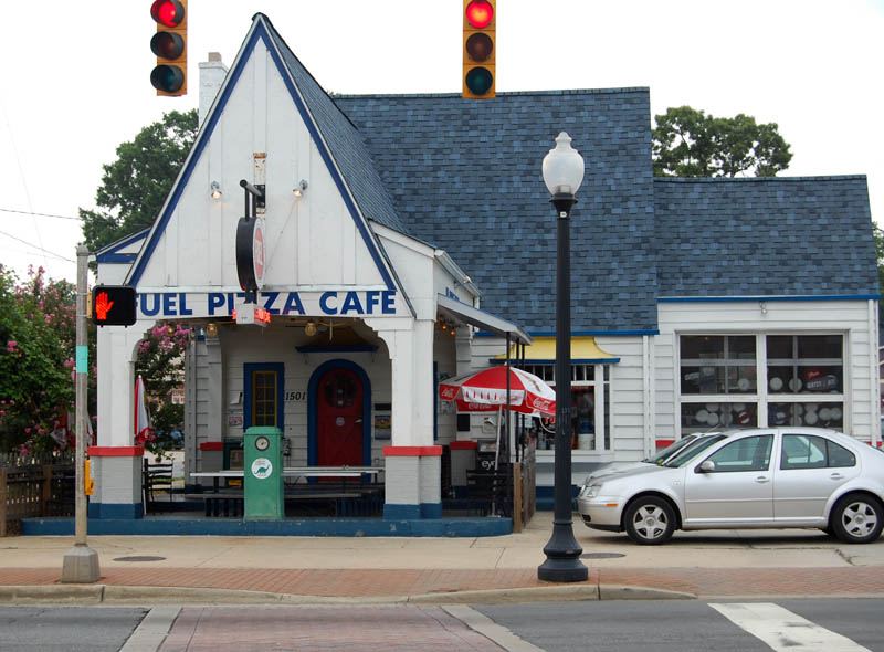 A former petrol station converted to a cafe: a small white weatherboard building with a steep blue shingle roof and a sharply pointed gable over the entrance porch, a red door, and a fascia sign reading 'Fuel Pizza Cafe'. An old petrol pump stands on the forecourt beside picnic benches and a parasol, with a silver car parked at the kerb and traffic signals overhead.