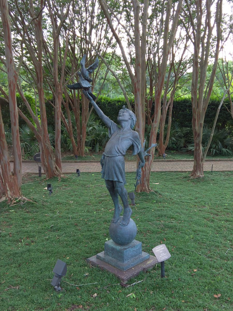 Bronze sculpture of a child standing on a sphere with one arm raised releasing a bird, on a stone plinth in front of crape myrtle trees.