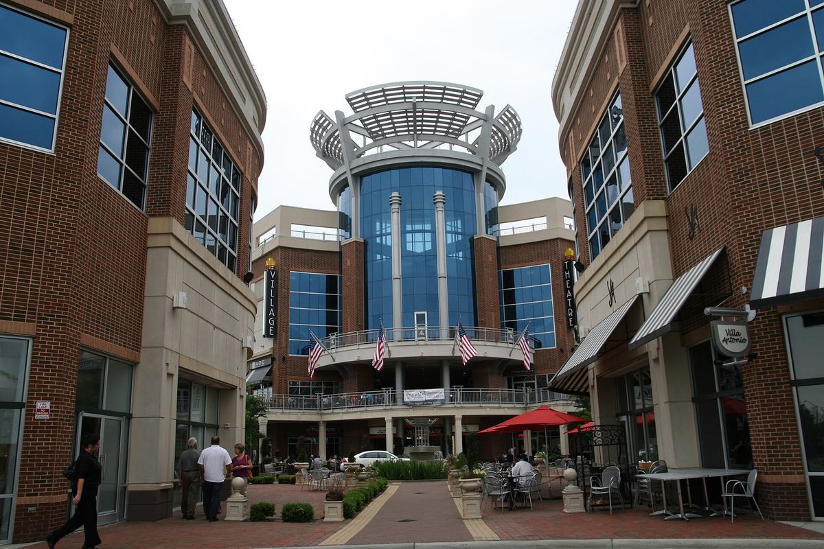 Retail and office development with a circular glass tower at the centre, canopied shopfronts, outdoor seating and people walking.